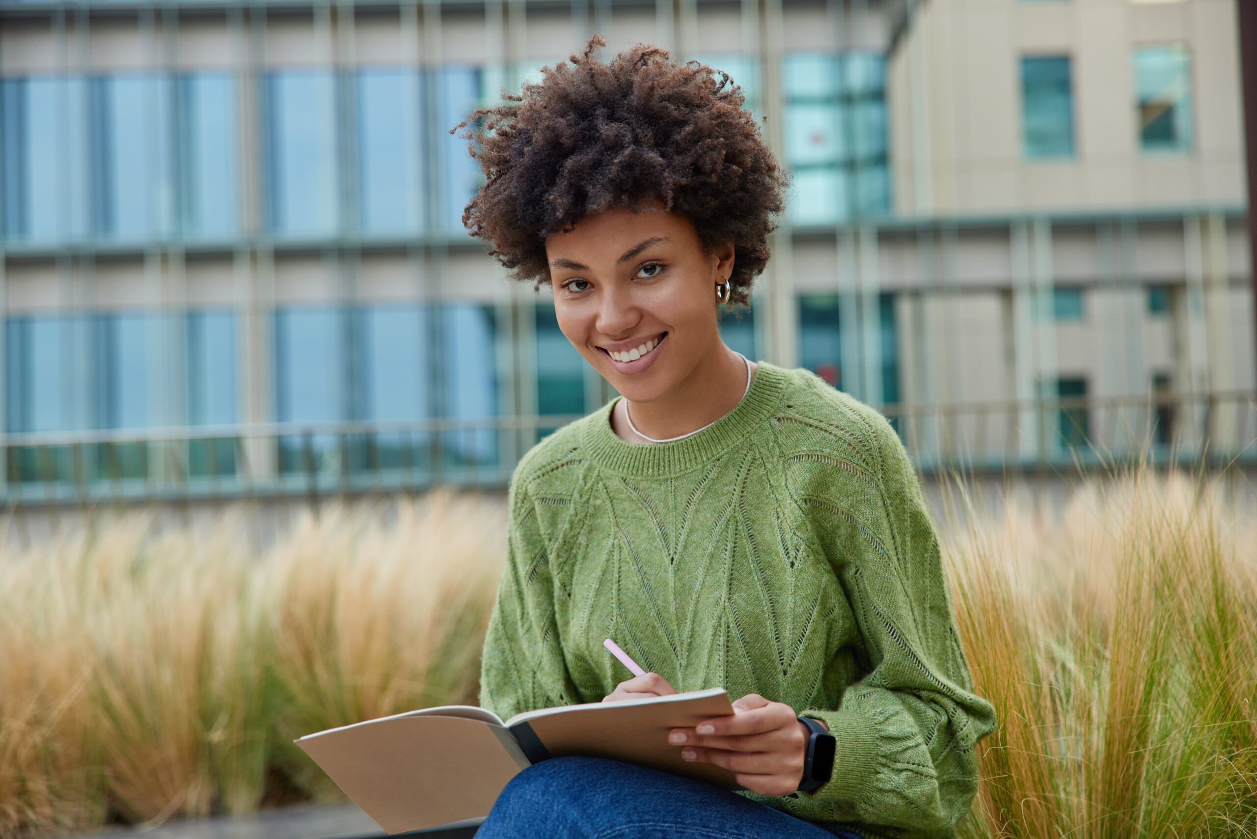 happy curly haired woman works writing essay puts down ideas notepad wears casual green jumper smiles positively camera poses outdoors against blurred backgroun